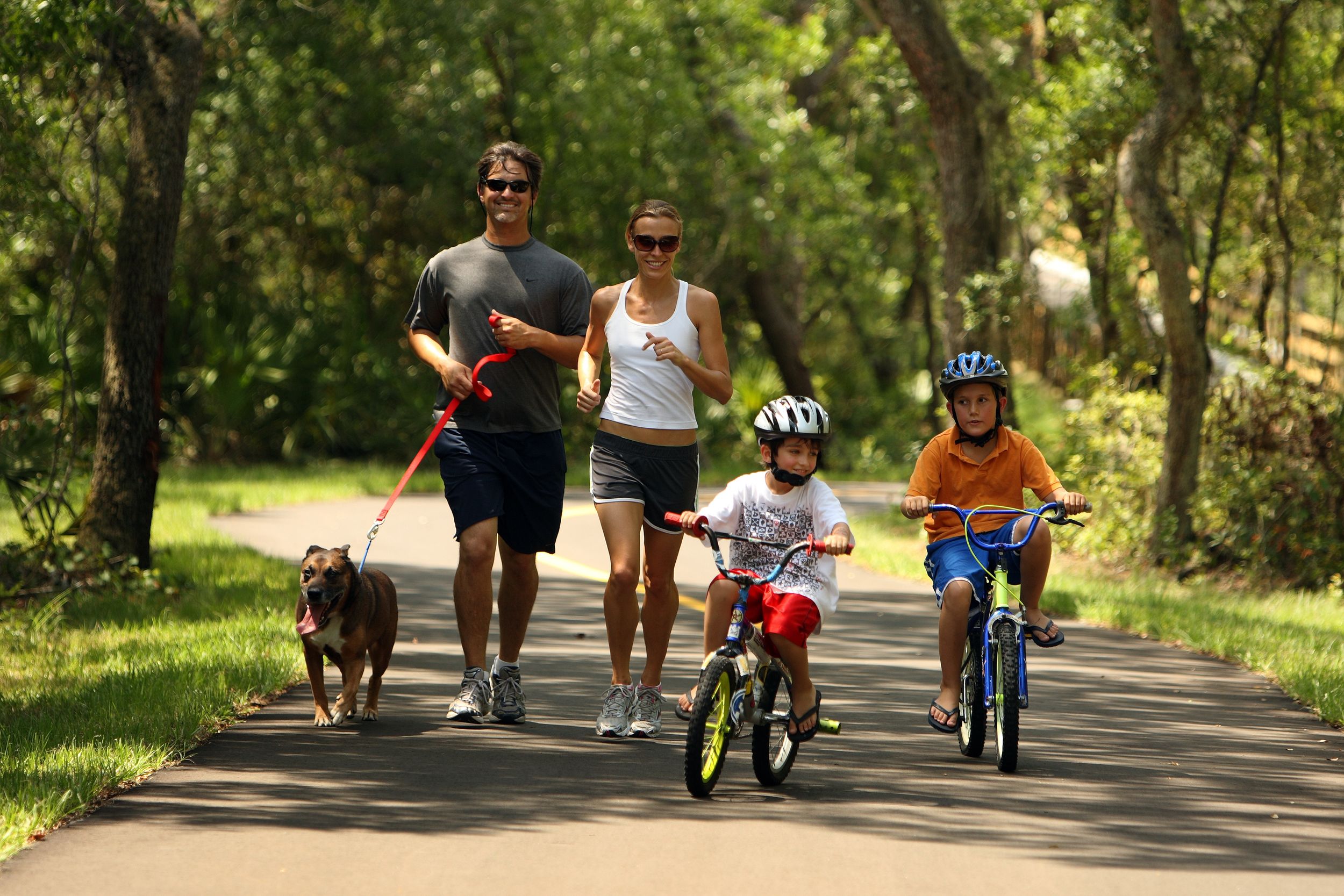 Parents running with dogs and children biking on a shady street.